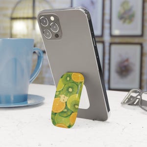 May include: A smartphone is propped up by a colorful phone grip. The grip features a pattern of green kiwi slices and orange citrus fruit. The phone is gray and the grip has a white stand.