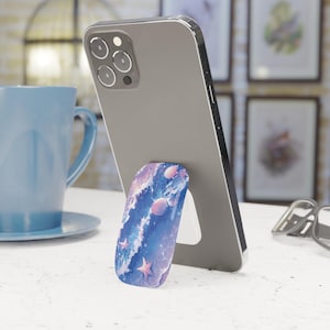 May include: A smartphone with a seashell and starfish design phone grip. The grip features a blue and purple watercolor design with pink seashells and starfish. The phone is gray and is propped up on a white surface.