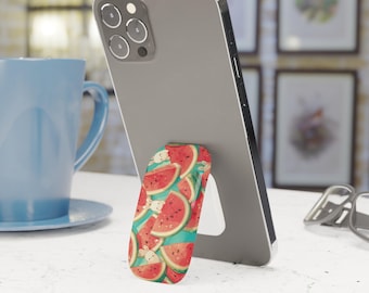 Può includere: Uno smartphone grigio è sostenuto da un supporto per telefono a tema anguria. Il supporto presenta fette di anguria rossa su sfondo turchese. Una tazza blu e opere d'arte incorniciate sono visibili.