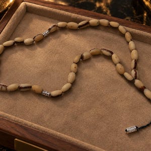 May include: A prayer bead strand featuring oval-shaped, cream-coloured beads with dark brown streaks. The strand is accented with two silver-toned metal spacers and terminates in a black cord with a silver-toned metal cylindrical end piece.