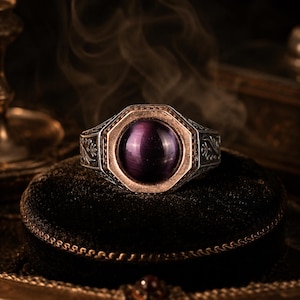 Könnte beinhalten: Ein silberner Ring mit einem runden, tiefvioletten Cabochon-Stein in einer achteckigen Fassung. Die Ringschiene ist an den Seiten mit detaillierten, eingravierten Blumenmustern verziert und weist ein kontrastierendes Metallfinish auf.