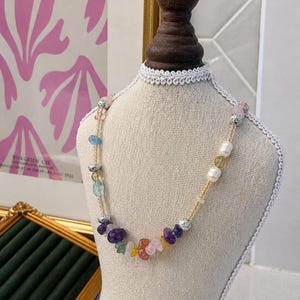 May include: A colorful gemstone necklace displayed on a mannequin. The necklace features a variety of stones in shades of purple, pink, yellow, and blue, interspersed with pearls and silver-colored beads. The necklace is strung on a delicate gold-colored chain.