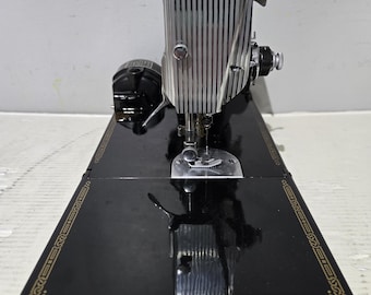 Puede incluir: Máquina de coser negra vintage con superficie brillante. La máquina presenta una cabeza plateada con líneas verticales y un borde dorado en los bordes de la base. Diseñada para coser, es un ejemplo clásico.