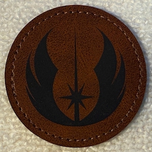 May include: A circular brown leather patch featuring a black Jedi Order symbol laser-engraved in the centre. The patch has a textured surface and is finished with a stitched border around the entire circumference of the circular shape.