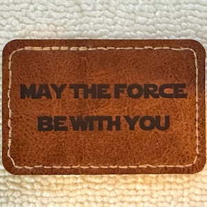 May include: A rectangular brown leather patch with rounded corners and white stitching along the edges. The text MAY THE FORCE BE WITH YOU is embossed in a dark, bold, sans-serif font across the center of the textured leather surface.