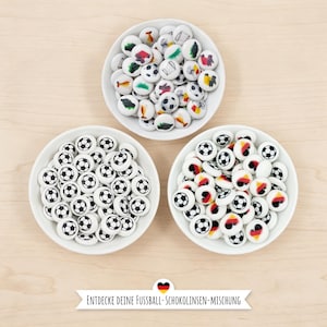 May include: Three white bowls filled with soccer ball-themed candies. Some candies feature soccer ball designs, while others have flags and drawings. The text "ENTDECKE DEINE FUSSBALL-SCHOKOLINSEN-MISCHUNG" is displayed on a banner.