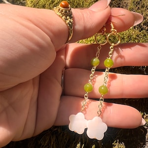 May include: A pair of dangle earrings with a gold chain and green beads. The earrings have a white flower charm at the bottom.