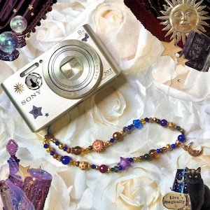 May include: A silver Sony digital camera with a Zeiss lens and 20.1 megapixels, decorated with small celestial stickers, positioned next to a beaded necklace featuring blue, purple, and gold beads, star and sun charms, and a crescent moon pendant.