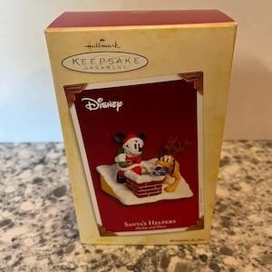 May include: A Hallmark Keepsake Ornament box featuring a Disney Santa's Helpers scene with Mickey Mouse in a Santa suit and Pluto wearing reindeer antlers by a brick chimney. The box displays the Hallmark logo, Disney branding, and text.