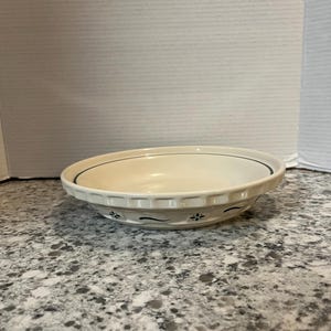 May include: Off-white ceramic pie dish with a slightly scalloped edge. The dish has a dark blue line around the rim and a repeating floral pattern in dark blue along the lower edge. The dish is empty and sits on a speckled surface.