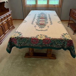May include: A rectangular tablecloth with a floral design. The tablecloth has a cream-colored background with a border of green, burgundy, and yellow flowers. The table is made of wood.