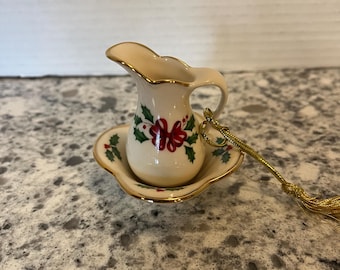May include: A miniature cream pitcher and matching saucer ornament featuring a cream-colored ceramic body with a painted holly and red bow pattern. The edges of the pitcher and saucer are trimmed in gold, and a gold hanging cord is attached.
