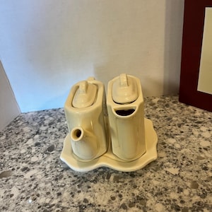 May include: A cream-colored ceramic cruet set consisting of two rectangular containers with matching lids, each featuring a small handle and spout, all resting together on a single, scalloped-edge tray that holds both pieces securely in place.