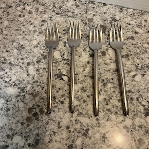 Puede incluir: Cuatro tenedores plateados con púas, dispuestos horizontalmente. Los tenedores tienen un diseño simple y clásico con mangos largos y delgados. Los tenedores están hechos de un metal brillante y reflectante, probablemente acero inoxidable.