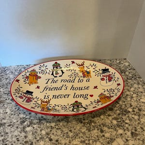 May include: An oval ceramic serving platter with a cream-colored base and a red rim. The center features the black text, "The road to a friend's house is never long," surrounded by illustrations of snowmen, reindeer, and penguins with lights.