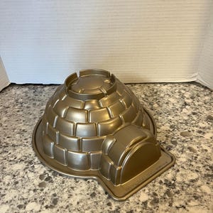 May include: A gold-colored igloo-shaped cake pan. The pan features a detailed brick pattern and a rounded doorway. The pan is made of metal and has a circular base.