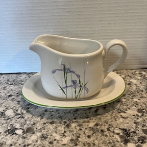 May include: White ceramic gravy boat with a matching saucer. The gravy boat features a handle and a spout for easy pouring. The set is decorated with a floral design of purple irises and green stems. The saucer has a green rim.