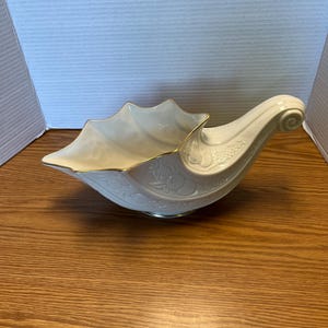 May include: A cream-colored ceramic cornucopia bowl featuring a scalloped rim with a gold-tone trim. The exterior surface is decorated with embossed fruit motifs, including pineapples and grapes, and the piece rests on a circular base.