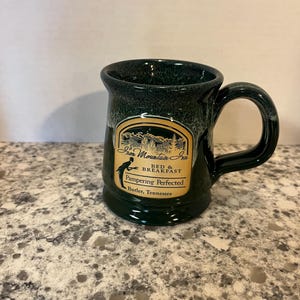 May include: A dark green ceramic mug with a speckled glaze finish. The front features a tan label with the text Iron Mountain Inn, Bed & Breakfast, Pampering Perfected, and Butler, Tennessee, along with a graphic of a person.