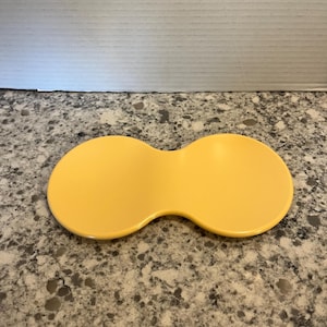 May include: A yellow, double-lobed, ceramic dish. The dish has a smooth, glossy finish and is sitting on a speckled gray countertop. The dish's shape is reminiscent of two connected circles.