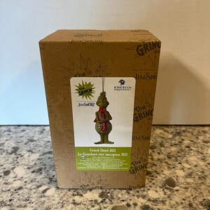 Pode incluir: Uma caixa de papelão marrom com a marca The Grinch e um rótulo mostrando um enfeite verde do Grinch usando um cachecol vermelho com o ano 2022. O rótulo inclui o texto The Grinch, Jim Shore, Enesco e Grinch Dated 2022.