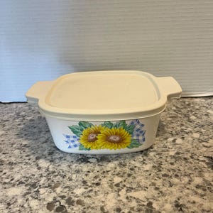 May include: Off-white rectangular food storage container with a matching lid. The container features a sunflower and blue gingham design. The lid has two handles. The container is made of plastic.
