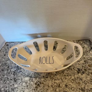Op de afbeelding: Crèmekleurige keramische broodmand, ovaal met uitgesneden ontwerp. Het woord "ROLLS" is zwart gedrukt. Voorzien van twee kleine handvatten. Geschikt voor het serveren van broodjes. Ongeveer 25 cm lang.