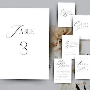 May include: A collection of elegant white wedding signs with black calligraphy. The signs include "Table 3", "Guest Book", "Cards + Gifts", "Welcome to our Wedding", "Bar Menu", and "Always in our Hearts". The signs are arranged against a soft, floral background.