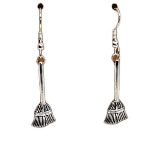 May include: A pair of silver-colored broom earrings. Each earring features a broom charm with a detailed bristle design and a hook for easy wear. The earrings are designed for a witchy or Halloween theme.