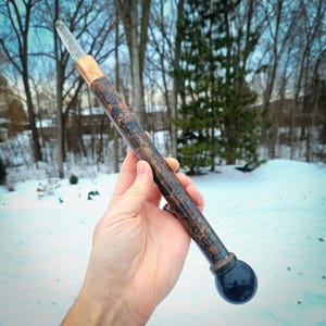 May include: A handcrafted wand with a clear crystal tip, copper accents, and a dark, textured wooden shaft. The wand has a black, spherical base and is held against a snowy backdrop.