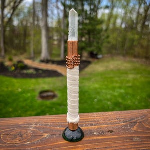 May include: A clear quartz crystal wand with copper accents. The wand features a clear crystal point, a copper tube, and wrapped copper wire. The handle is wrapped in white fabric and sits on a dark green base.