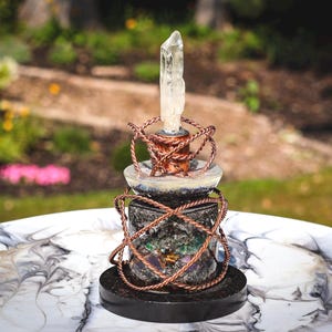 May include: A decorative object featuring a clear, pointed quartz crystal set atop a dark, textured base. The structure is wrapped in twisted copper wire and includes a smooth, light-colored disc element, all resting on a solid black circular stand.
