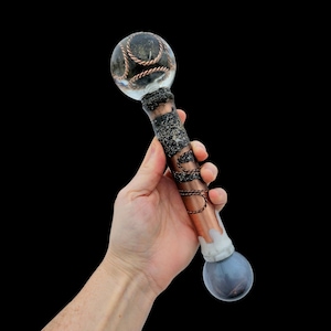 May include: A handcrafted wand with a clear orb top, featuring copper wire accents. The handle is copper-colored with dark textured detailing and a gray and white base. The wand is held against a black background.