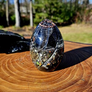 May include: A clear, egg-shaped resin art piece with a copper wire top. The interior features a mix of dark and iridescent materials. The piece sits on a wooden surface, with a blurred outdoor background.