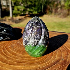 May include: A purple and green crystal orb with embedded dried flowers and herbs. The orb is made of resin and has a smooth, polished surface.