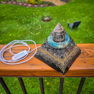 May include: A pyramid-shaped object made of resin containing dark stone fragments, gold-colored metallic flakes, and a central blue-tinted section wrapped in copper wire. A white power cord with a four-button control switch is attached to the base.