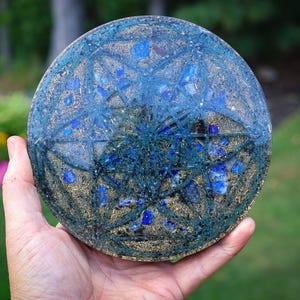 May include: A circular resin disc featuring a geometric flower of life pattern in dark blue. The surface is embedded with small, vibrant blue lapis lazuli stone fragments and fine gold-colored metallic shavings, creating a textured, layered appearance.