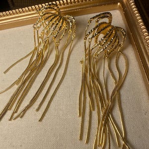 Peut inclure: Une paire de boucles d'oreilles pendantes dorées composées d'un groupe d'arches métalliques texturées et incurvées sur le dessus, d'où pendent plusieurs longues chaînes serpent flexibles, créant un effet de frange en cascade poli.