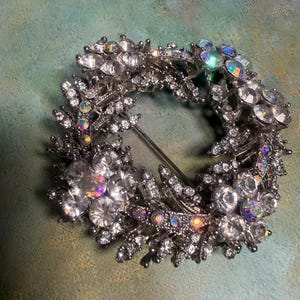 May include: A circular brooch featuring a silver-toned metal frame adorned with clear, faceted rhinestones and iridescent crystals. The design incorporates floral motifs and leafy sprigs arranged in a wreath shape, with a pin mechanism visible.
