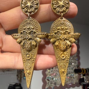 May include: A pair of gold-toned drop earrings featuring a circular stud with a starburst pattern connected to a triangular pendant. The pendant displays a detailed embossed bee at the centre, surrounded by intricate geometric and floral textures.