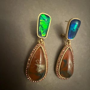 May include: A pair of gold-toned drop earrings. Each earring features a rectangular iridescent top in green or blue, and a teardrop-shaped bottom with a brown, white, and blue pattern, framed by pink crystals.