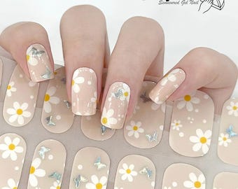 May include: Nail polish wraps with a daisy and butterfly design. The wraps are beige with white daisies and silver butterflies. The wraps are shown on a hand with a light pink nail polish.