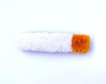 Könnte beinhalten: Eine weiße, rechteckige Haarspange mit flauschiger Textur, die einer Zigarette ähnelt. Das Ende der Spange ist ein Quadrat aus orangefarbenem Garn. Die Spange ist vor einem weißen Hintergrund platziert.