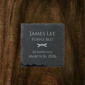 Op de afbeelding: Een vierkante, donkergrijze leistenen onderzetter met gegraveerde tekst. De tekst luidt "JAMES LEE", "PURPLE BELT", een gestileerde riemafbeelding, "BJJ AMERICANA" en "MARCH 16, 2026". De onderzetter ligt op een donkere houten ondergrond.