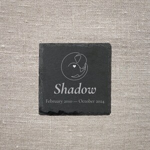 Pode incluir: Uma base para copos quadrada de ard&oacute;sia preta com um desenho a tra&ccedil;o branco de um c&atilde;o e um cora&ccedil;&atilde;o. O nome "Shadow" est&aacute; abaixo do desenho, com as datas "Fevereiro 2010 - Outubro 2024".