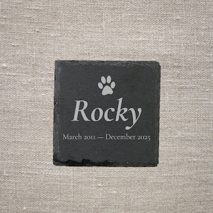 Pode incluir: Base quadrada de ard&oacute;sia preta com uma pegada e o nome "Rocky" em branco. O texto "Mar&ccedil;o 2011 - Dezembro 2025" est&aacute; abaixo. Um item de mem&oacute;ria.