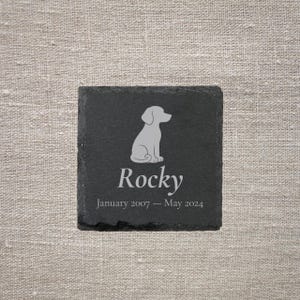 Pode incluir: Uma base para copos quadrada de ard&oacute;sia preta com uma silhueta cinza claro de um c&atilde;o e o nome "Rocky". Abaixo do nome est&atilde;o as datas "Janeiro 2007 - Maio 2024". A base para copos est&aacute; sobre um fundo de linho neutro.