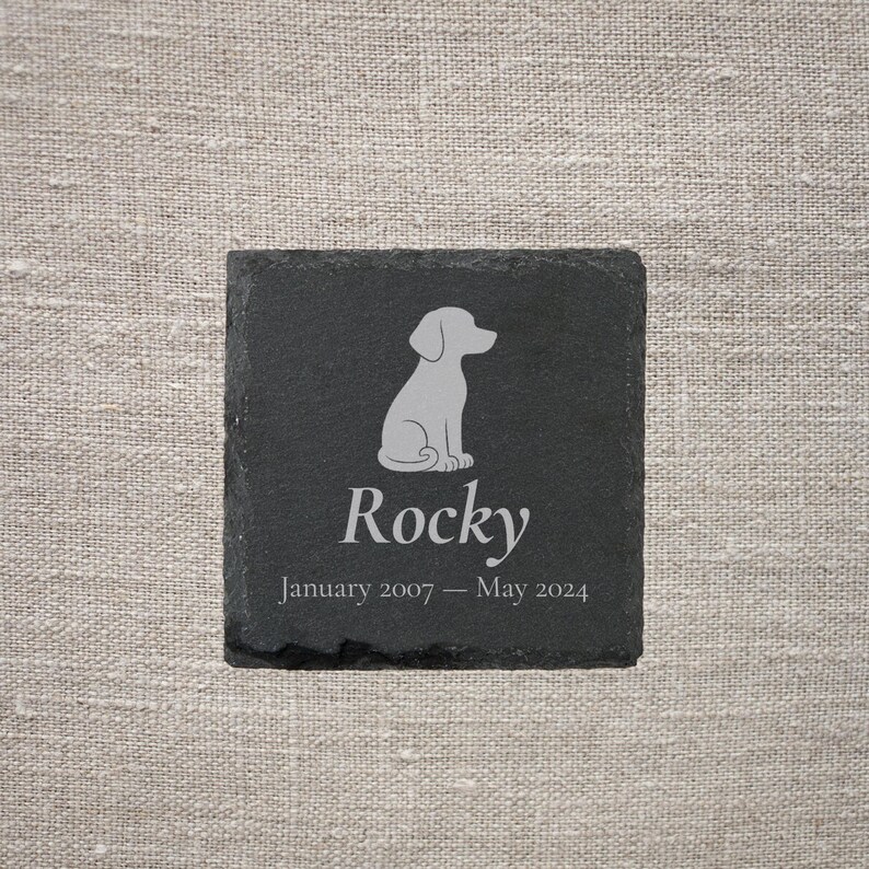 Pode incluir: Uma base para copos quadrada de ard&oacute;sia preta com uma silhueta cinza claro de um c&atilde;o e o nome "Rocky". Abaixo do nome est&atilde;o as datas "Janeiro 2007 - Maio 2024". A base para copos est&aacute; sobre um fundo de linho neutro.