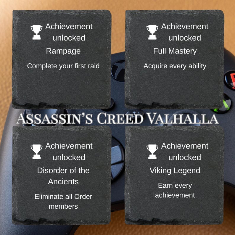 Puede incluir: Cuatro posavasos cuadrados de pizarra negra con texto blanco e iconos de trofeos. Cada posavasos muestra un logro del videojuego Assassin's Creed Valhalla. El mando de la consola es visible en el fondo.