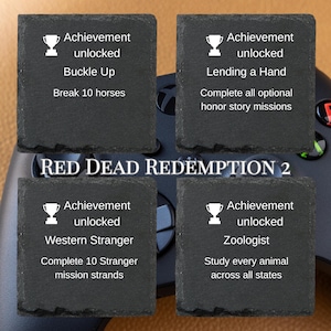 Puede incluir: Cuatro posavasos cuadrados de pizarra con texto blanco y gr&aacute;ficos de trofeos. Cada posavasos muestra un logro del videojuego Red Dead Redemption 2. Los logros incluyen "Buckle Up", "Lending a Hand", "Western Stranger" y "Zoologist".